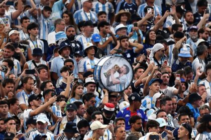 Large cheering crowd in blue-and-white jerseys; a person holds a round drum featuring a kissing couple at center.