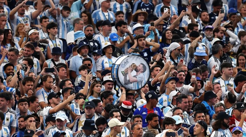 Large cheering crowd in blue-and-white jerseys; a person holds a round drum featuring a kissing couple at center.