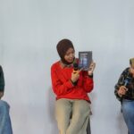 Three women sit on stools in a panel discussion; the middle woman in a red sweater holds a book and speaks into a microphone while the others listen.