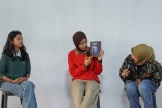 Three women sit on stools in a panel discussion; the middle woman in a red sweater holds a book and speaks into a microphone while the others listen.