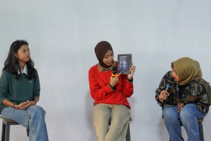 Three women sit on stools in a panel discussion; the middle woman in a red sweater holds a book and speaks into a microphone while the others listen.