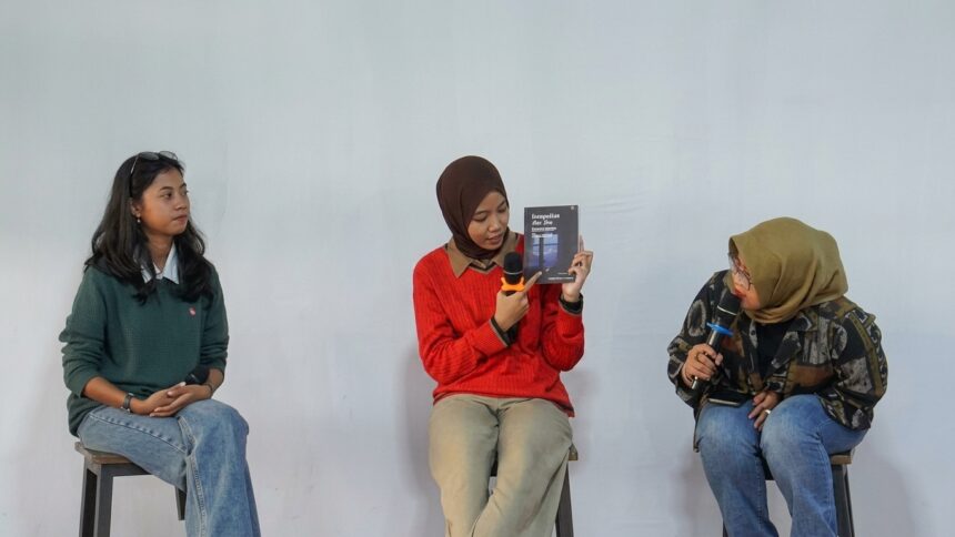 Three women sit on stools in a panel discussion; the middle woman in a red sweater holds a book and speaks into a microphone while the others listen.