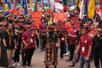 Street festival scene with a performer in ornate ceremonial armor at center, surrounded by a crowd of marchers and colorful flags and banners.