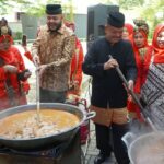 People in traditional red outfits cook around two large outdoor pots, with men stirring a soup over open flames.