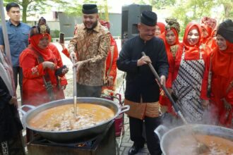 People in traditional red outfits cook around two large outdoor pots, with men stirring a soup over open flames.