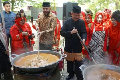 People in traditional red outfits cook around two large outdoor pots, with men stirring a soup over open flames.
