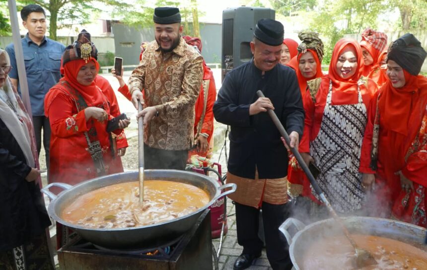 People in traditional red outfits cook around two large outdoor pots, with men stirring a soup over open flames.