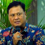 Man in a blue floral shirt speaks into a handheld microphone with green plants in the background