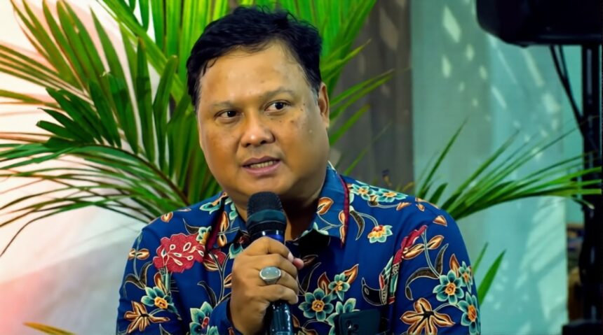 Man in a blue floral shirt speaks into a handheld microphone with green plants in the background