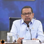 Man in a light-blue shirt speaks at a government press conference, seated behind a table with a microphone and a nameplate that reads 'KEPALA BADAN KOMUNIKASI PEMERINTAH'