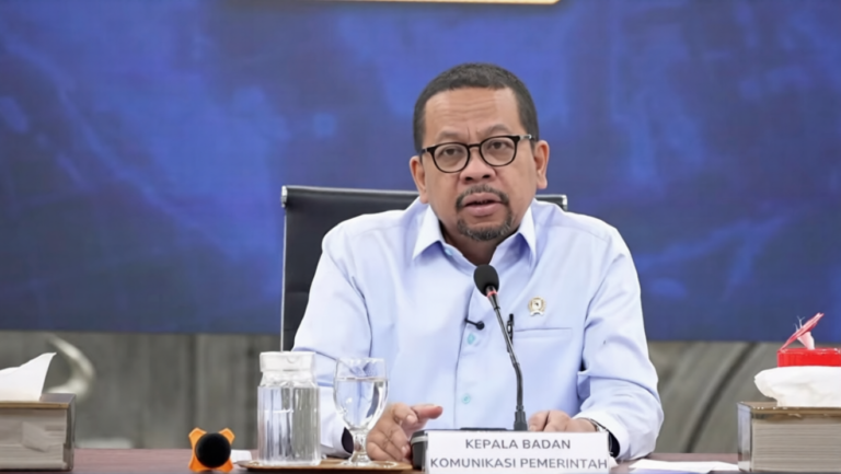 Man in a light-blue shirt speaks at a government press conference, seated behind a table with a microphone and a nameplate that reads 'KEPALA BADAN KOMUNIKASI PEMERINTAH'