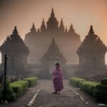 Person in a pink plaid wrap stands on a path before an ancient temple with tall, spire-topped towers in mist.