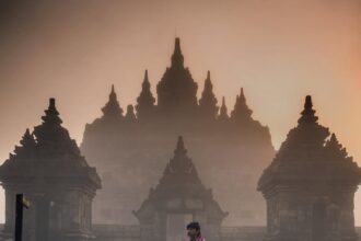Person in a pink plaid wrap stands on a path before an ancient temple with tall, spire-topped towers in mist.