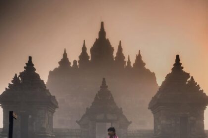 Person in a pink plaid wrap stands on a path before an ancient temple with tall, spire-topped towers in mist.