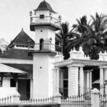 Black-and-white photo of a colonial-style building with a small tower, gated entrance, and palm trees in the background.
