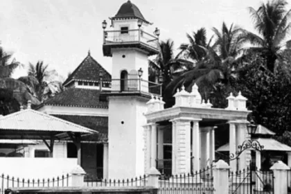 Black-and-white photo of a colonial-style building with a small tower, gated entrance, and palm trees in the background.