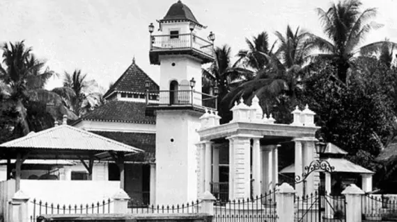 Black-and-white photo of a colonial-style building with a small tower, gated entrance, and palm trees in the background.