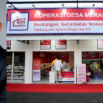 Close-up of a man in a suit with glasses on the left, next to a large red and white banner for KOPERASI DESA MERAH PUTIH.