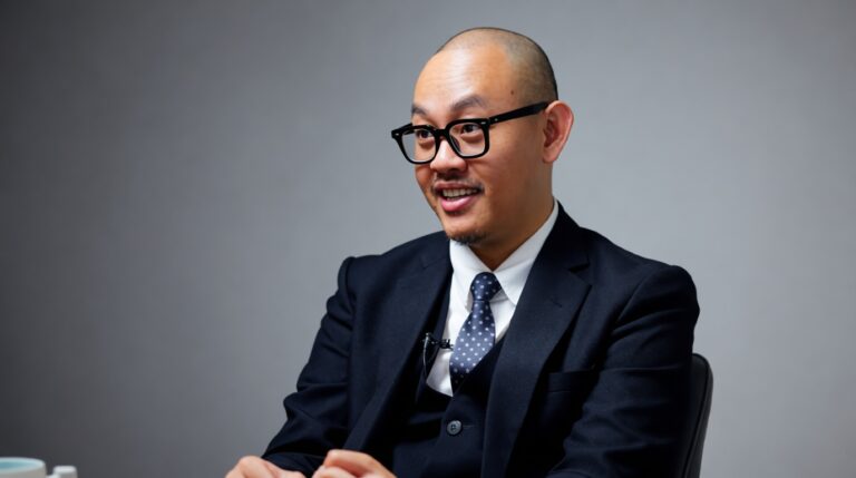 Man in a dark suit and polka-dot tie sits in an interview setting, speaking with a smile and a lapel mic visible on his jacket