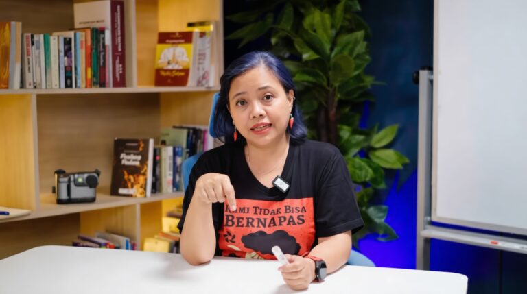 Woman with blue hair sitting at a white table, pointing at the camera, holding a marker; shelves with books behind her.