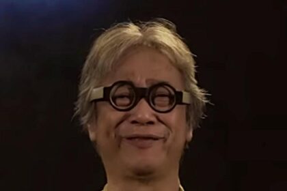 Person with short blond hair and round glasses, wearing a yellow-green shirt, smiling at the camera against a dark background.