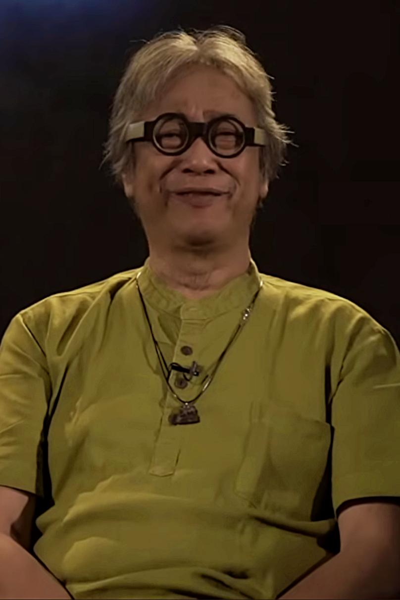 Person with short blond hair and round glasses, wearing a yellow-green shirt, smiling at the camera against a dark background.