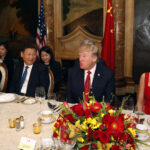 Diplomatic dinner scene with formally dressed attendees seated along a table, a large red‑yellow flower centerpiece, and flags in the background.