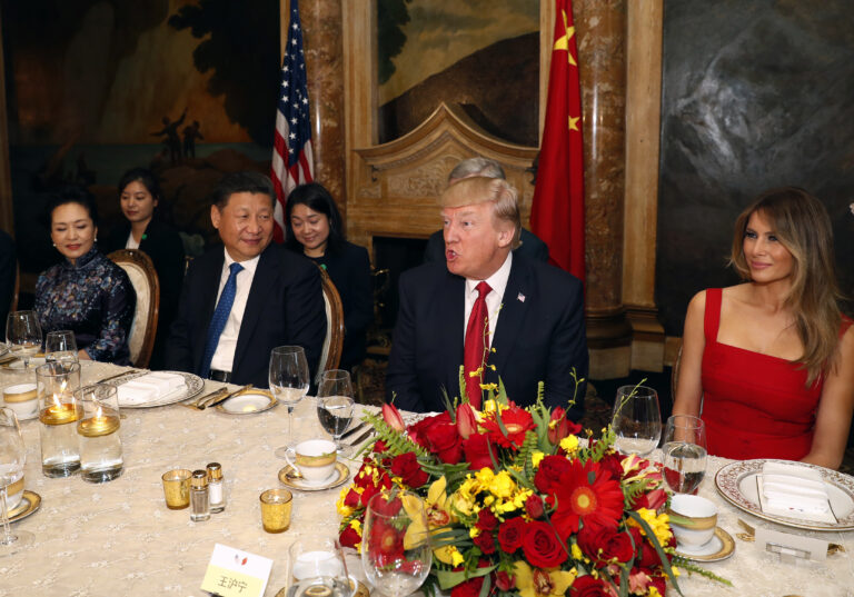 Diplomatic dinner scene with formally dressed attendees seated along a table, a large red‑yellow flower centerpiece, and flags in the background.