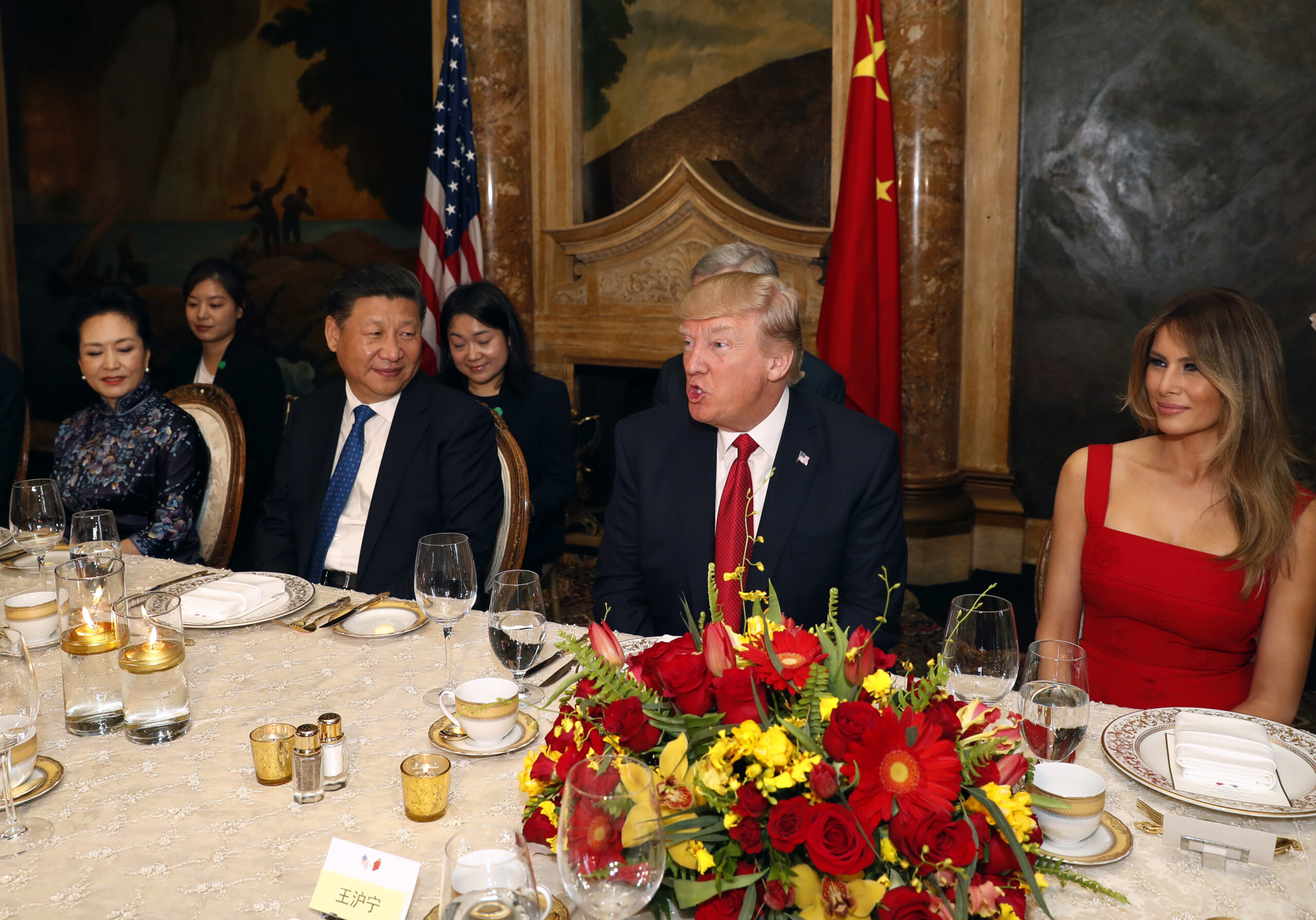 Diplomatic dinner scene with formally dressed attendees seated along a table, a large red‑yellow flower centerpiece, and flags in the background.