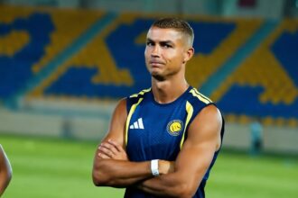 Athlete in a blue sleeveless jersey with yellow accents stands on a field with arms crossed, stadium seats blurred in the background.