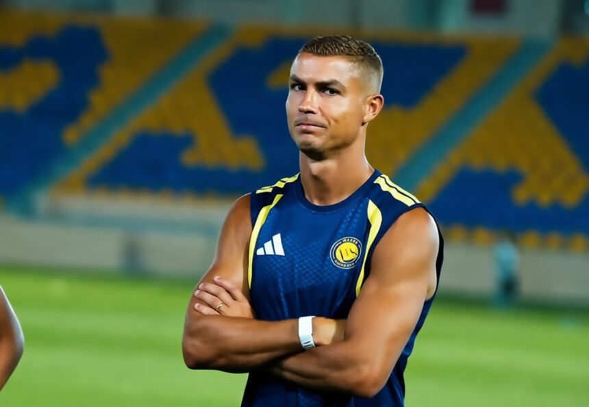 Athlete in a blue sleeveless jersey with yellow accents stands on a field with arms crossed, stadium seats blurred in the background.