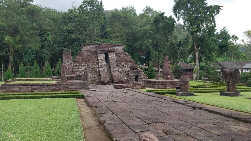 Ancient stone temple with a stepped doorway, surrounded by trimmed lawns and tall trees.