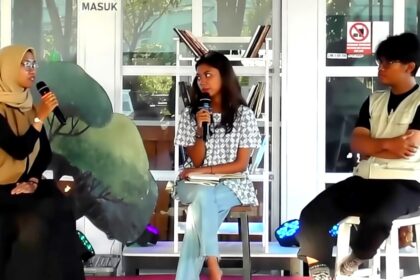 Three people seated on a panel: a hijab-wearing woman on the left speaks into a microphone, a woman in a patterned top sits center with a notebook, and a man in a light vest sits on the right in front of glass doors and shelves.