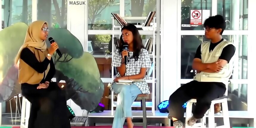 Three people seated on a panel: a hijab-wearing woman on the left speaks into a microphone, a woman in a patterned top sits center with a notebook, and a man in a light vest sits on the right in front of glass doors and shelves.