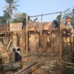 Group of construction workers on wooden scaffolding building a brick wall on a single-story house, outdoors with palm trees in the background