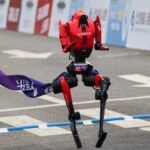 Red, HONOR-branded humanoid racing robot lifting one leg at a finish-line banner on a paved road track.