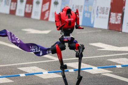 Red, HONOR-branded humanoid racing robot lifting one leg at a finish-line banner on a paved road track.