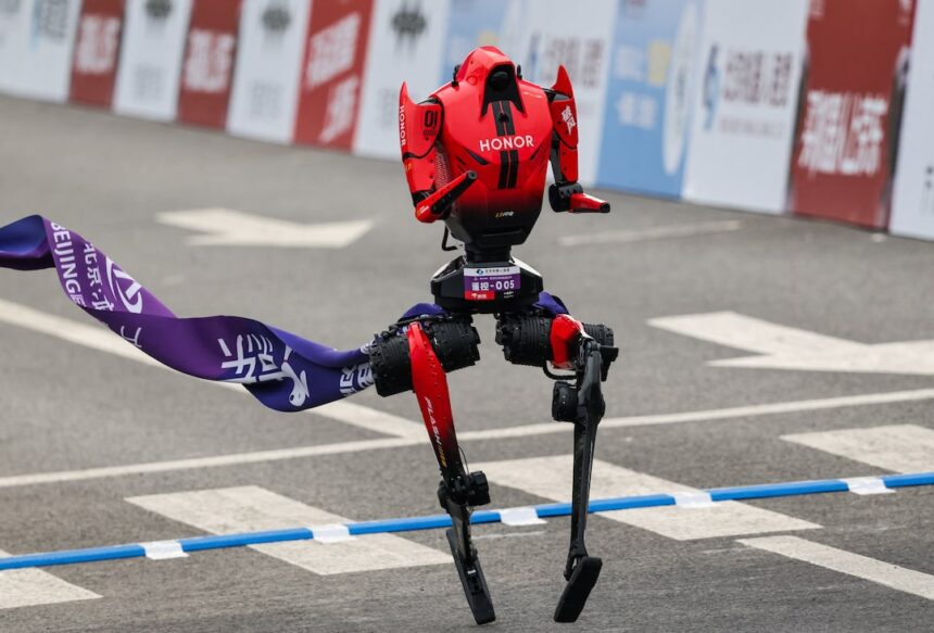Red, HONOR-branded humanoid racing robot lifting one leg at a finish-line banner on a paved road track.