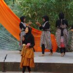 Group of women in matching patterned batik dresses and hijabs on an outdoor stage with an orange drape in the background, preparing for a performance, microphones on the edge.