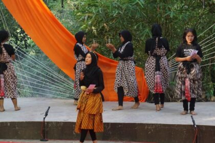 Group of women in matching patterned batik dresses and hijabs on an outdoor stage with an orange drape in the background, preparing for a performance, microphones on the edge.