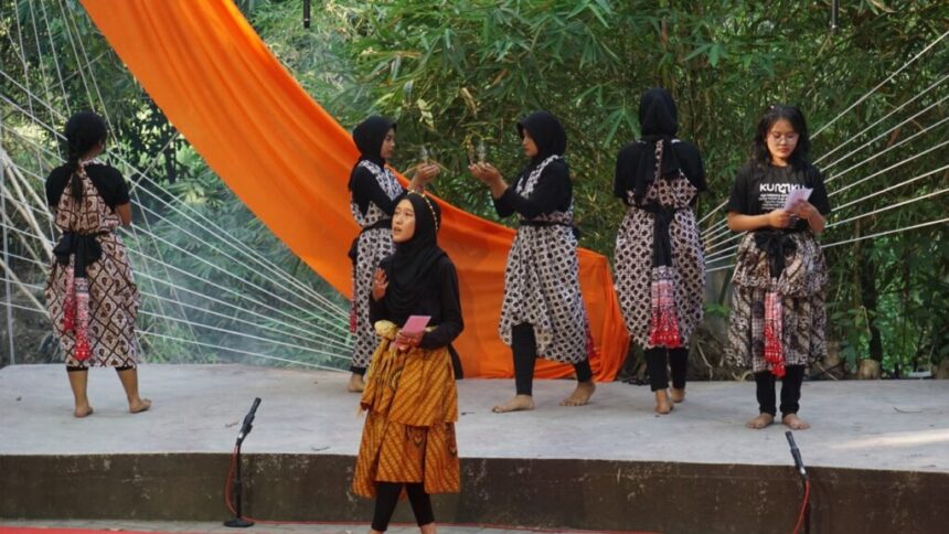 Group of women in matching patterned batik dresses and hijabs on an outdoor stage with an orange drape in the background, preparing for a performance, microphones on the edge.