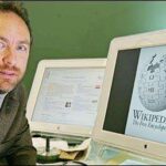 Man sits at a desk with two computer monitors; one displays the Wikimedia globe logo (Wikipedia).