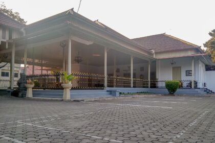 Open-air pavilion with white columns and gold-trimmed railing, connected to a small white building, set beside a paved parking area.