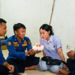 Four people in blue uniforms sit on the floor as one passes a cake with candles reading '22' to a woman, celebrating a birthday indoors.