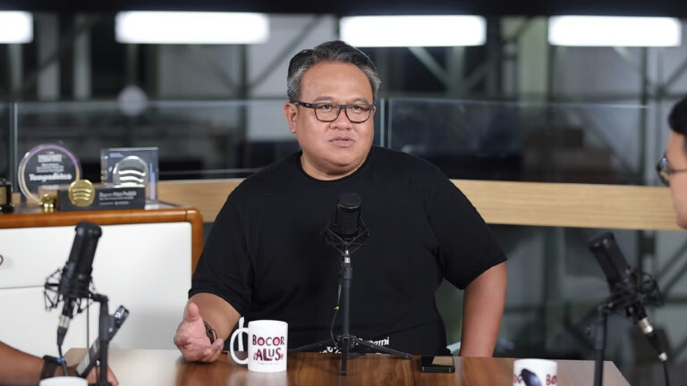 Man in a black t-shirt speaks into a studio microphone at a podcast setup, with mugs on the table in the foreground and awards visible in the background.