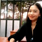Smiling woman with dark hair in a black top sitting at a wooden table indoors near a glass door with trees outside.