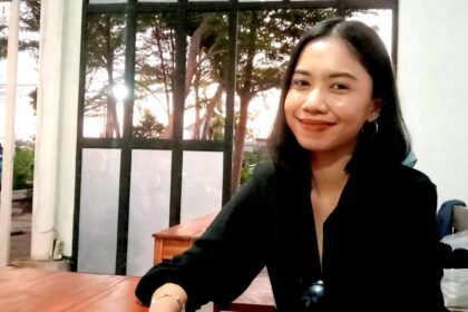 Smiling woman with dark hair in a black top sitting at a wooden table indoors near a glass door with trees outside.