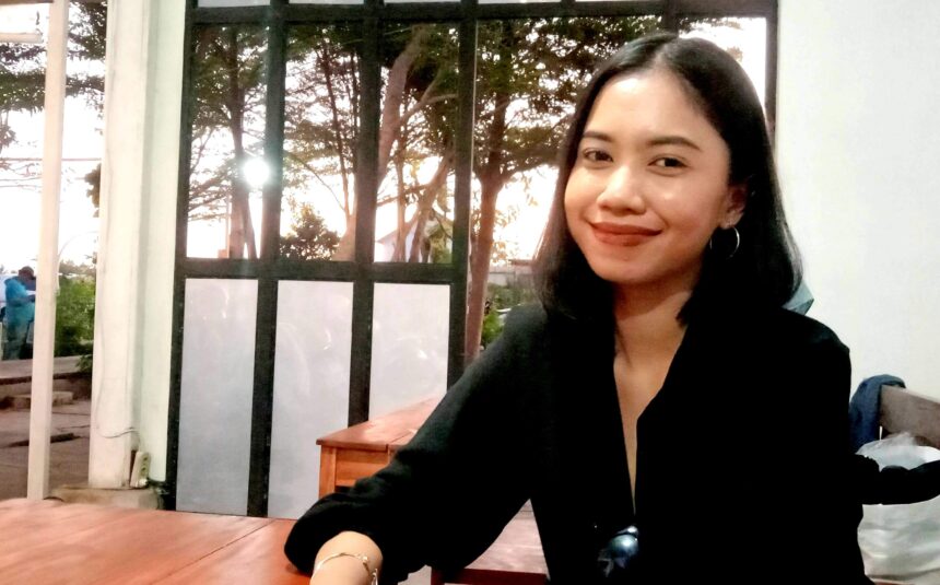 Smiling woman with dark hair in a black top sitting at a wooden table indoors near a glass door with trees outside.