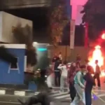 Nighttime street protest with a large fire and smoke in the background; people move around debris.