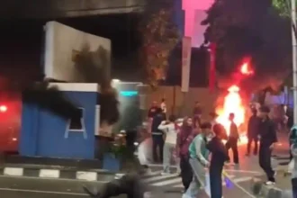 Nighttime street protest with a large fire and smoke in the background; people move around debris.